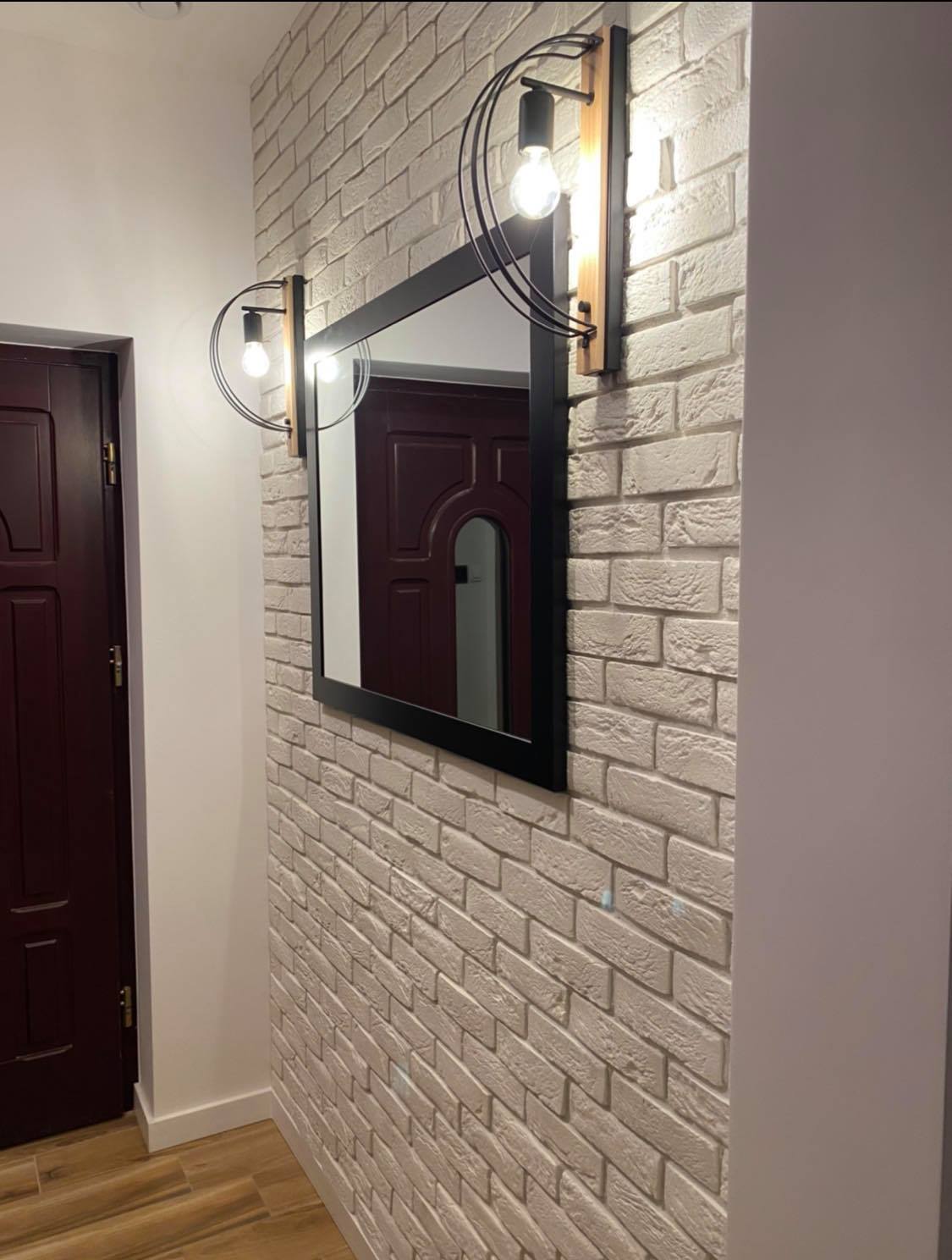 Ściana z dekoracyjnej cegły w odcieniach bieli z zamontowanym lustrem w czarnej ramie i nowoczesnymi lampami ściennymi z drewnianymi i metalowymi elementami, odbicie drzwi w lustrze.