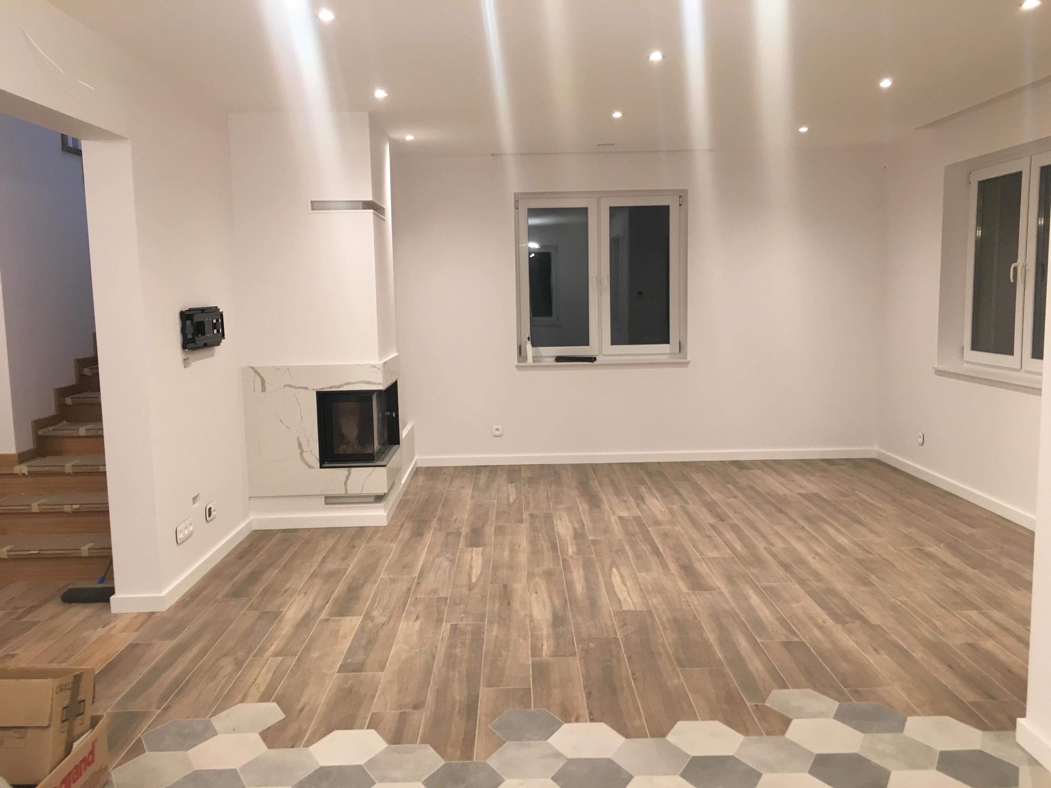 Salon z nową drewnopodobną podłogą, marmurowym kominkiem i częściowo ułożoną podłogą heksagonalną w odcieniach szarości, widoczne schody w tle.