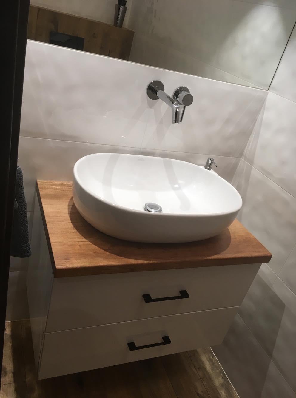 Nowoczesna łazienka z owalną, białą umywalką nablatową na szafce z drewnianym blatem i białymi frontami z czarnymi uchwytami, bateria umywalkowa ścienna w kolorze chromu, białe płytki na ścianie.