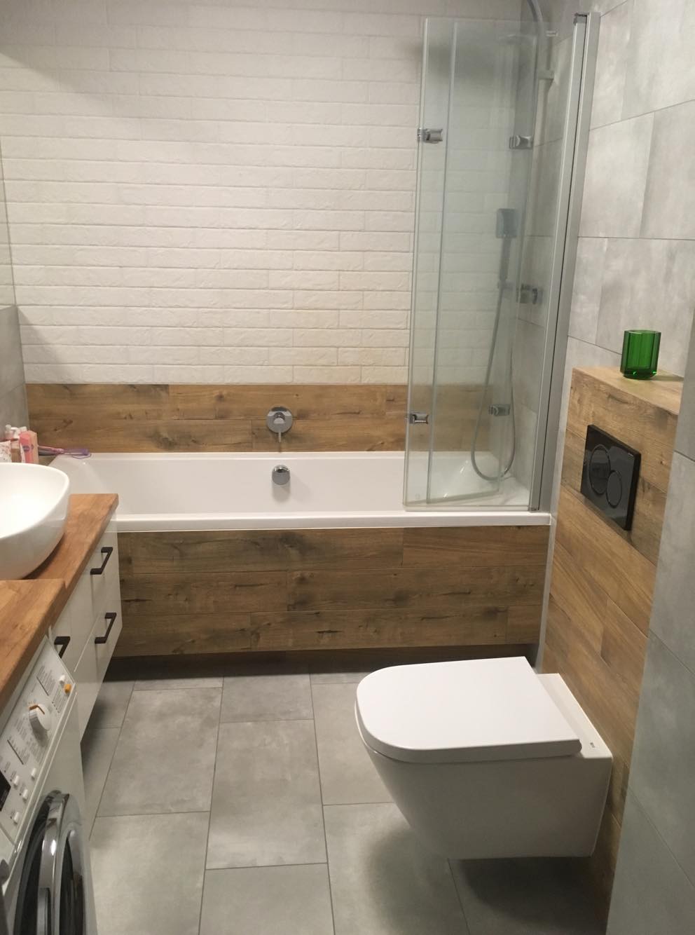 Nowoczesna łazienka z wanną obudowaną drewnem, kabiną prysznicową ze szklanymi drzwiami, podwieszaną toaletą i umywalką na blacie z pralką.