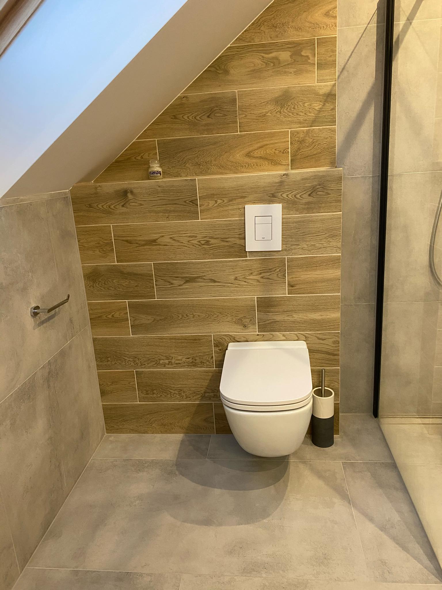 Nowoczesna toaleta wisząca w łazience na poddaszu, wykończonej płytkami imitującymi drewno i kamień, z widoczną spłuczką podtynkową i akcesoriami łazienkowymi.