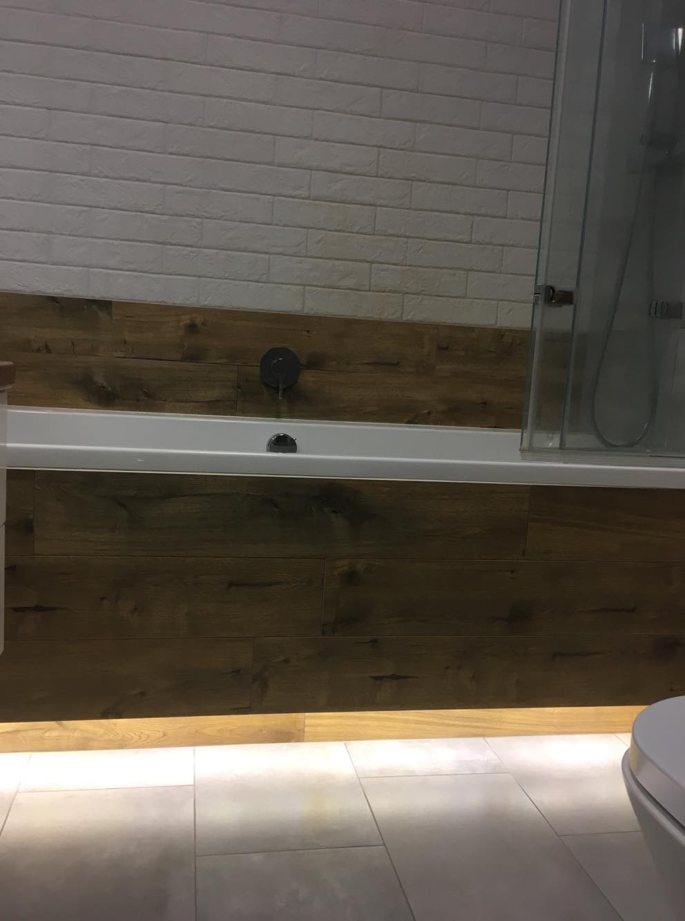 Nowoczesna łazienka z białą cegłą na ścianie, drewnopodobną obudową wanny z podświetleniem LED i szklanym panelem prysznicowym.