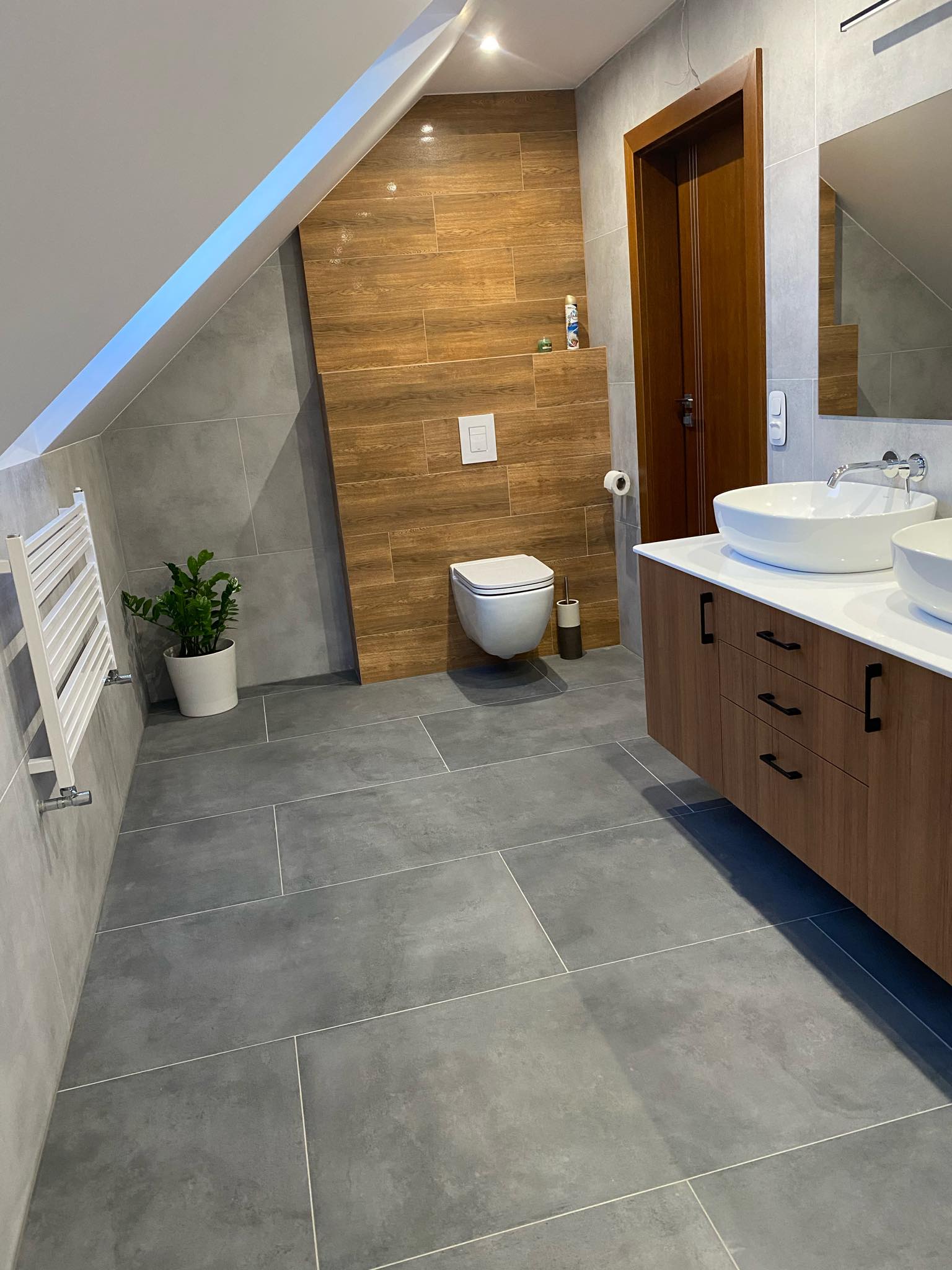 Nowoczesna łazienka na poddaszu z szarymi płytkami podłogowymi i ściennymi, drewnopodobną ścianą za toaletą, białymi umywalkami nablatowymi, brązową szafką pod umywalkami i grzejnikiem drabinkowym.