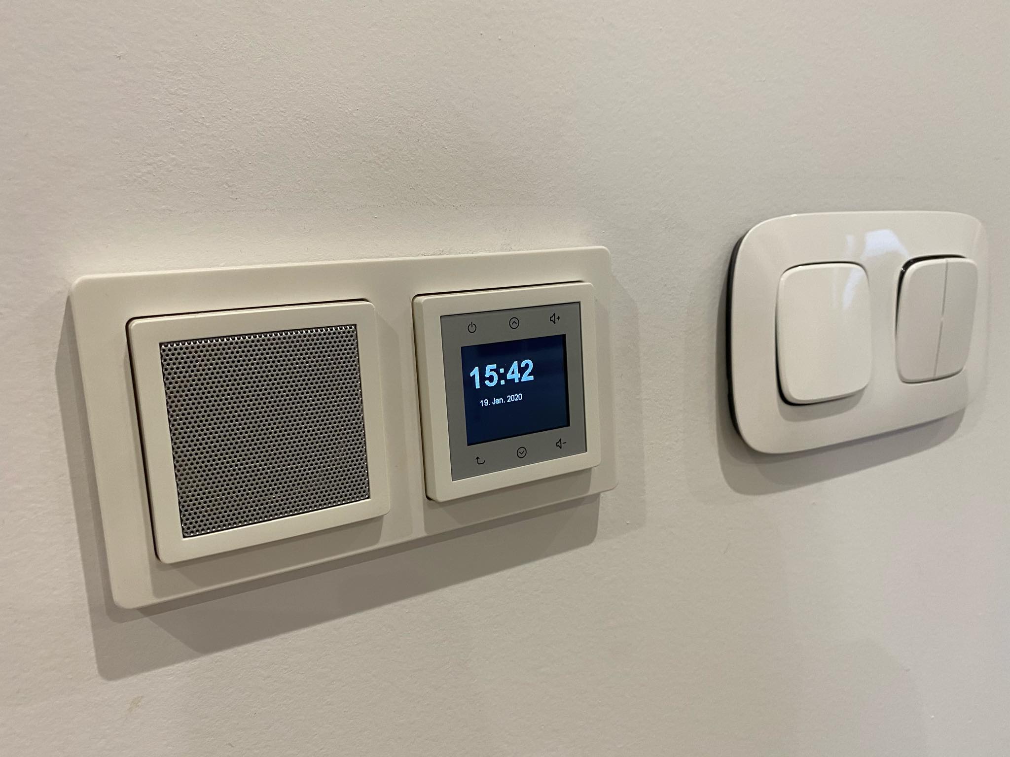 Panel sterowania inteligentnym domem z wyświetlaczem pokazującym godzinę i datę, zamontowany na ścianie obok podwójnego włącznika światła.