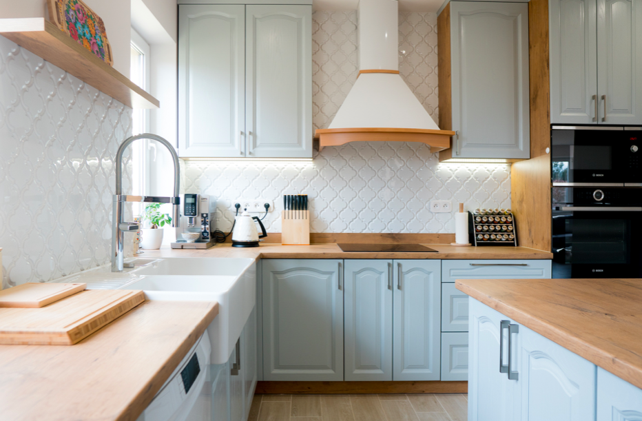 Jasna kuchnia w stylu retro z szarymi szafkami, drewnianymi blatami i białymi płytkami ściennymi w marokański wzór, widok na zlew, ekspres do kawy i płytę indukcyjną.