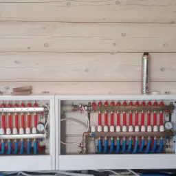 Rozdzielacz ogrzewania podłogowego zamontowany w białej skrzynce, widoczne rury czerwone i niebieskie, manometry, oraz drewniana ściana w tle.