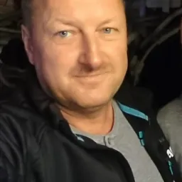 Portret mężczyzny o niebieskich oczach, ubranego w szarą koszulkę i czarną bluzę z niebieskimi akcentami, na tle ciemnego wnętrza z elementami instalacji.