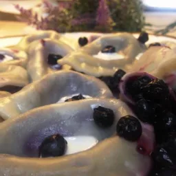 Delikatne pierogi z jagodami polane sosem jagodowym i kleksem śmietany, podane na białym talerzu z lawendą w tle.