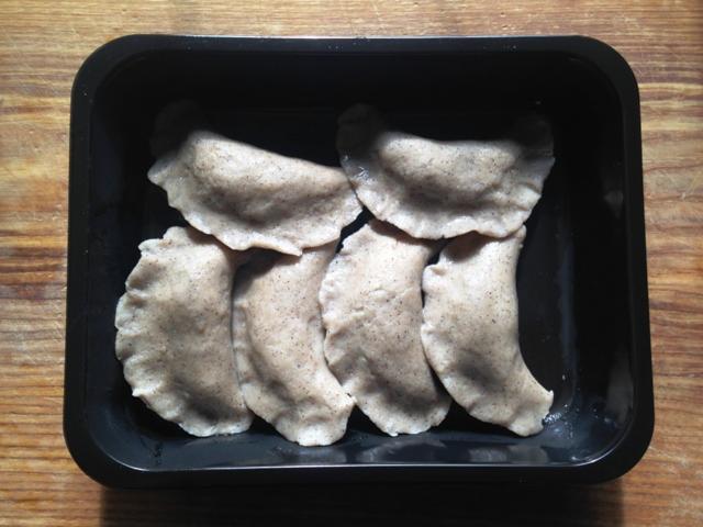 Siedem pierogów z ciemnego ciasta, ułożonych w czarnym, plastikowym pojemniku na tle drewnianego blatu.