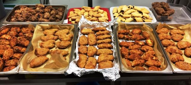 Rzędy metalowych tac z różnorodnymi wyrobami garmażeryjnymi, w tym krokiety na folii aluminiowej, ciasteczka z nadzieniem oraz smażone kotlety, ustawione na blacie roboczym.