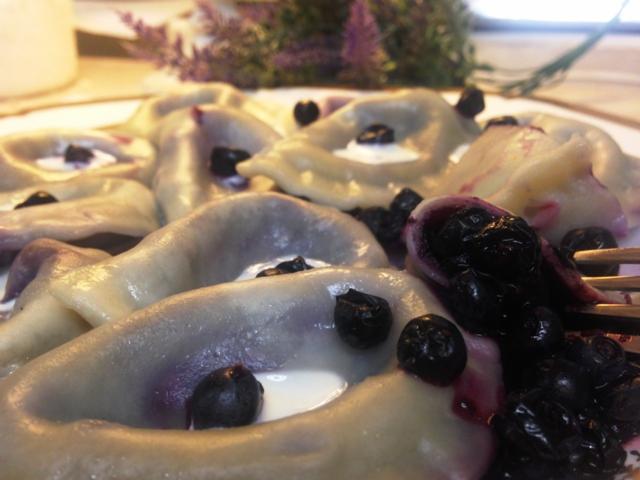 Delikatne pierogi z jagodami polane sosem jagodowym i kleksem śmietany, podane na białym talerzu z lawendą w tle.