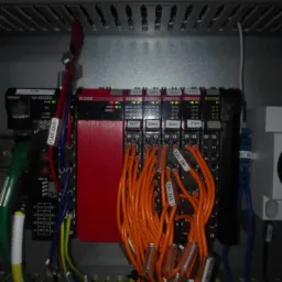 Panel sterowania z modułami Omron, widoczne liczne kable, w tym pomarańczowe z oznaczeniami 20A1 XA3 i 20A1 XB1, oraz moduły SL3300, SID800, SOD400, OD4256, umieszczone w szarej szafie sterowniczej...