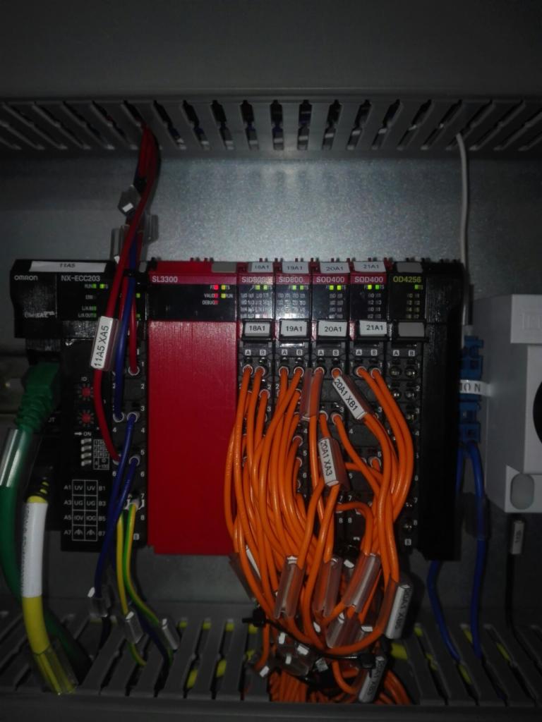 Panel sterowania z modułami Omron, widoczne liczne kable, w tym pomarańczowe z oznaczeniami 20A1 XA3 i 20A1 XB1, oraz moduły SL3300, SID800, SOD400, OD4256, umieszczone w szarej szafie sterowniczej...
