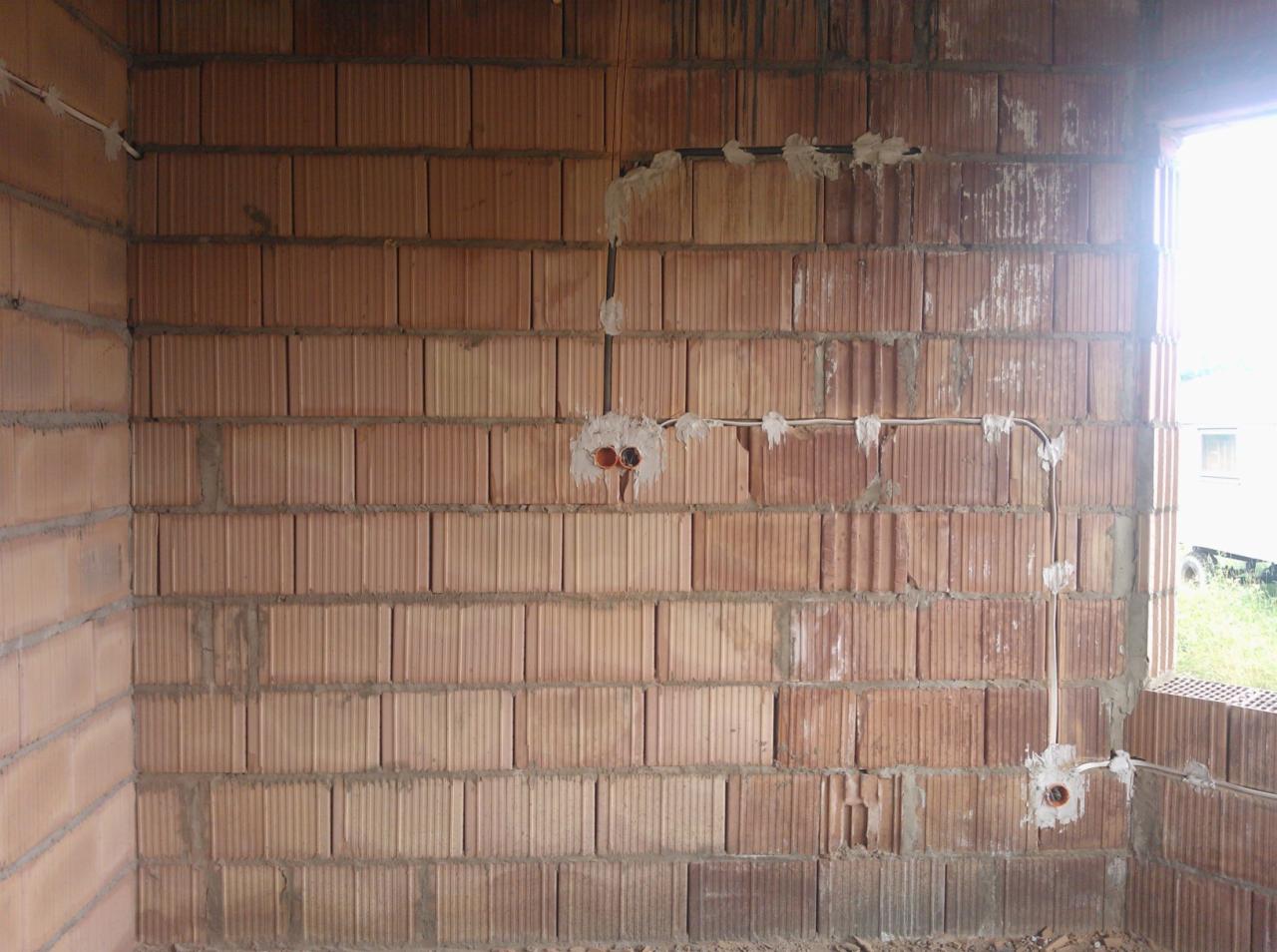 Instalacja elektryczna natynkowa w surowym budynku z cegły: przewody poprowadzone po ścianie i puszki podtynkowe zamocowane gipsem.