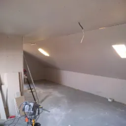 Poddasze w trakcie wykańczania: ściany obłożone płytami gipsowo-kartonowymi, widoczne łączenia, wystający kabel, resztki materiałów budowlanych na podłodze, drabina i narzędzia w tle.