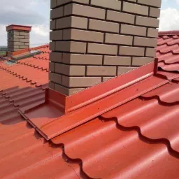 Czerwona blachodachówka z ceglanym kominem i obróbkami blacharskimi w kolorze dachu, widok z góry.