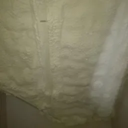 Nierówna, jasna warstwa izolacji natryskowej na poddaszu, z widocznymi śladami po rozpyleniu i fragmentem niezabezpieczonej rury.