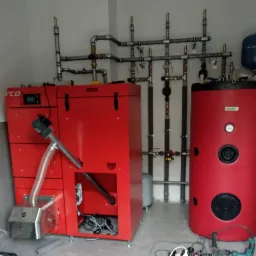 Czerwony piec na pellet z zasobnikiem ciepłej wody, widoczne rury i instalacja, ułożone na szarych płytkach, w tle białe ściany, przewody elektryczne na podłodze.