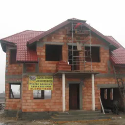 Niedokończony, dwukondygnacyjny dom z czerwonej cegły z częściowo położonym czerwonym dachem, rusztowaniem i drabiną, na tle pochmurnego nieba i błotnistego terenu.