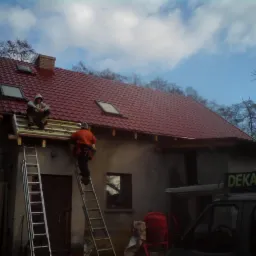 Dekarze montują dachówkę na dachu budynku z oknami dachowymi, jeden siedzi na dachu, drugi wchodzi po drabinie, widoczny samochód z napisem 'Dekarstwo'.
