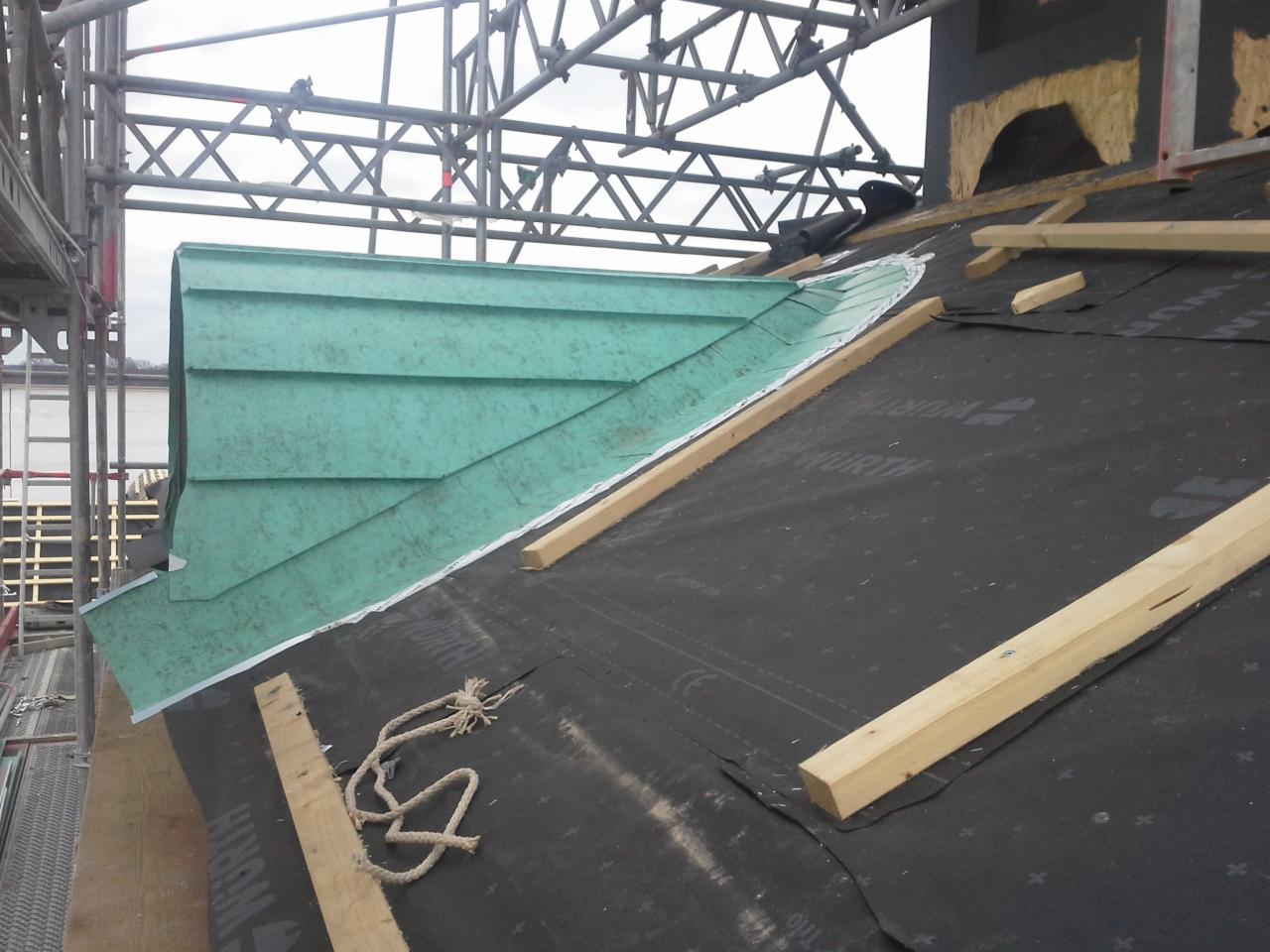 Montaż miedzianej lukarny na dachu pokrytym membraną, widoczne drewniane łaty i fragment rusztowania.