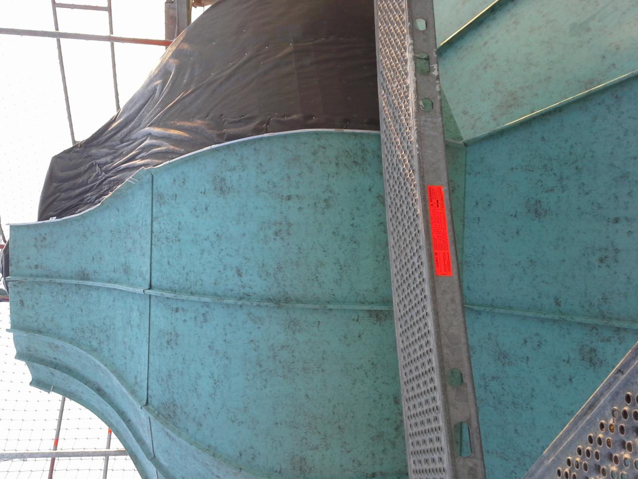 Fragment dachu z blachy trapezowej o nietypowym, zakrzywionym kształcie, częściowo zakryty czarną folią ochronną, widoczne rusztowanie i drabina z metalu.