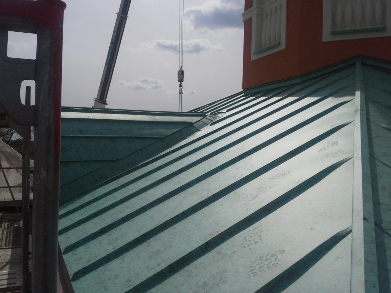 Nowe, miedziane pokrycie dachowe z widocznymi śladami butów, w tle żuraw budowlany i fragment pomarańczowej elewacji budynku z oknami.