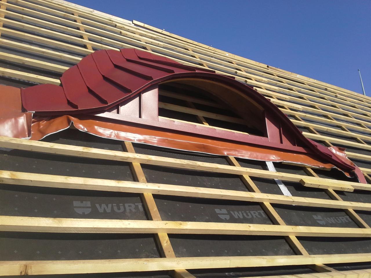 Widok z dołu na konstrukcję dachu z oknem lukarną w trakcie montażu, widoczne drewniane łaty, czarna membrana i fragment czerwonej blachy na oknie.