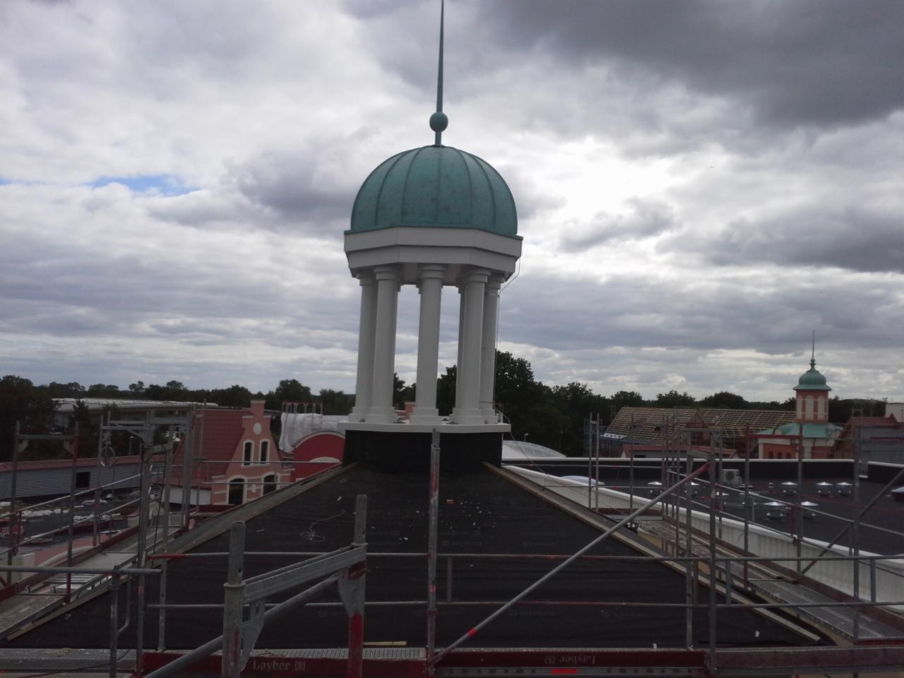 Widok z góry na dach pokryty czarną papą, z centralnie umieszczoną wieżyczką z zieloną kopułą i białymi kolumnami, otoczoną rusztowaniem Layher na tle pochmurnego nieba.