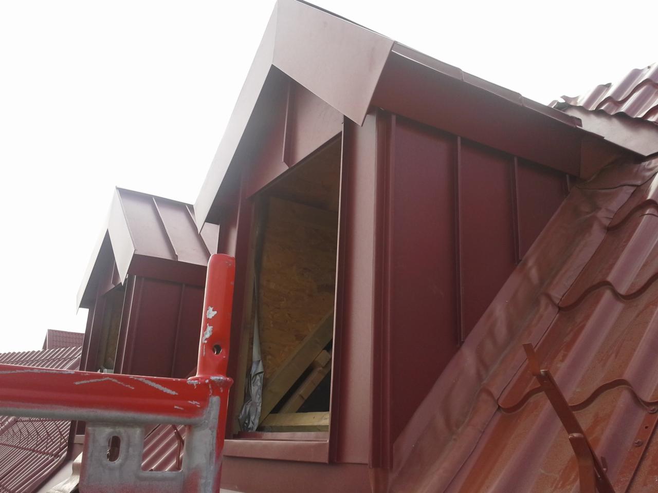 Czerwona blacha na dachu podczas budowy lukarny, widoczne rusztowanie i fragment istniejącego pokrycia dachowego w kolorze ceglastym.