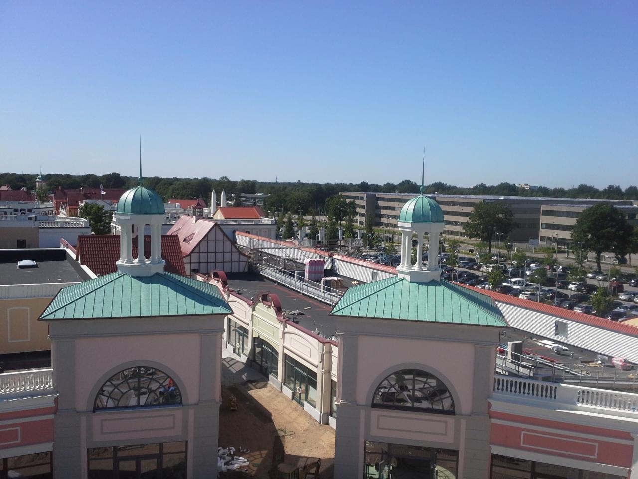 Widok z góry na budynki handlowe z charakterystycznymi wieżyczkami zwieńczonymi zielonymi kopułami, w tle parking i budynki biurowe.