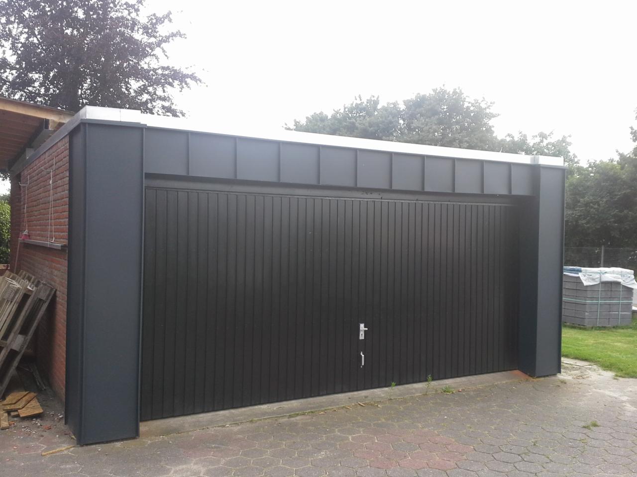 Nowoczesny garaż z płaskim dachem i ciemnoszarą blachą trapezową na elewacji, widoczne obróbki blacharskie na łączeniu dachu ze ścianą, paleta i kostka brukowa w tle.