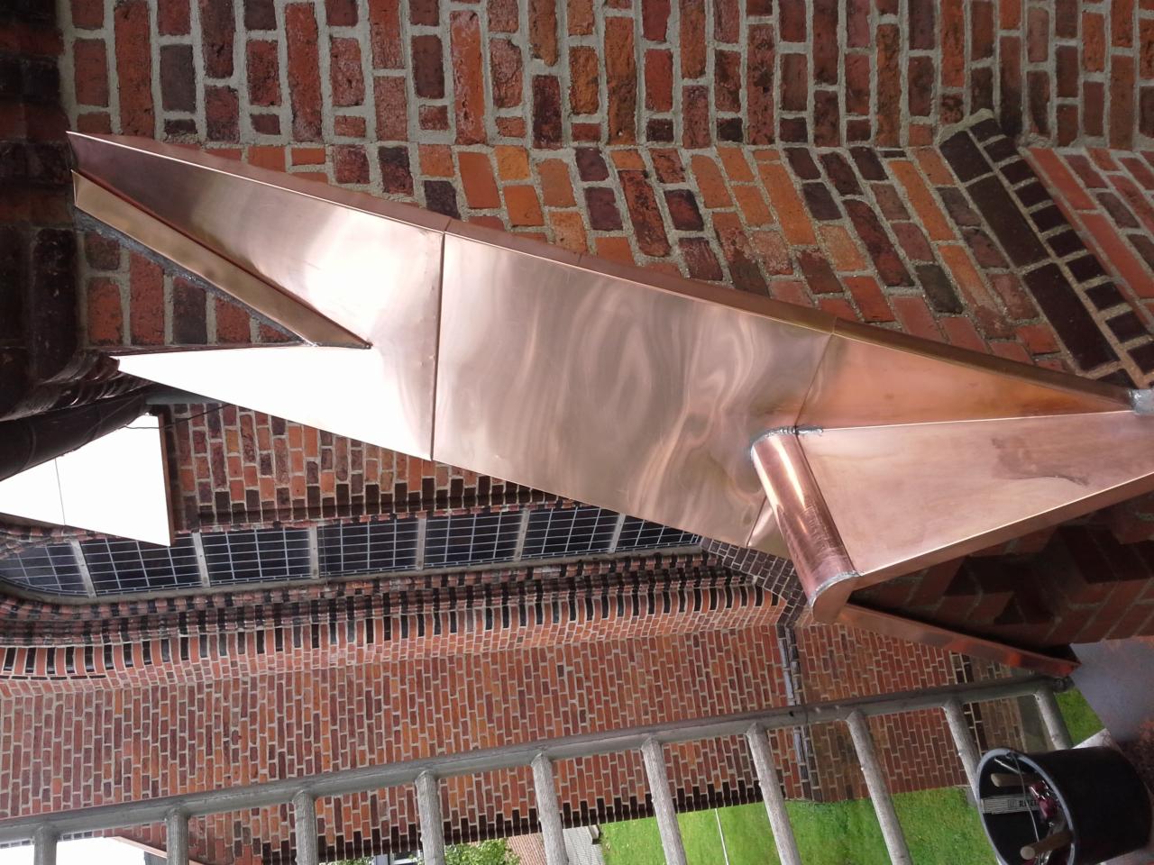 Miedziane elementy wykończeniowe dachu na tle ceglanej ściany, widoczne łączenia blachy i fragment rynny.