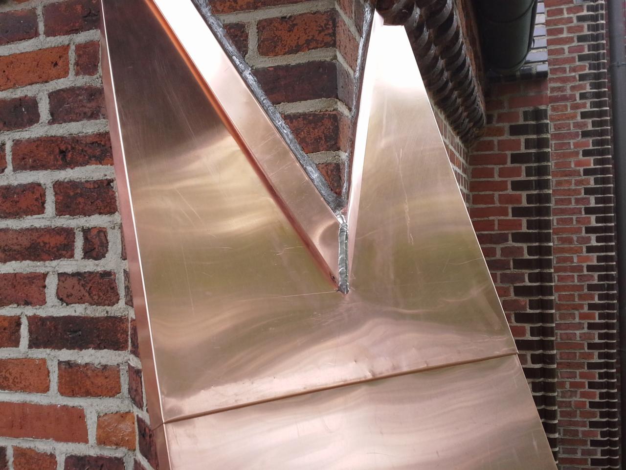 Miedziana obróbka blacharska na styku dachu z ceglanym kominem, widoczne spawy i ślady obróbki na metalu, fragment rynny w tle.