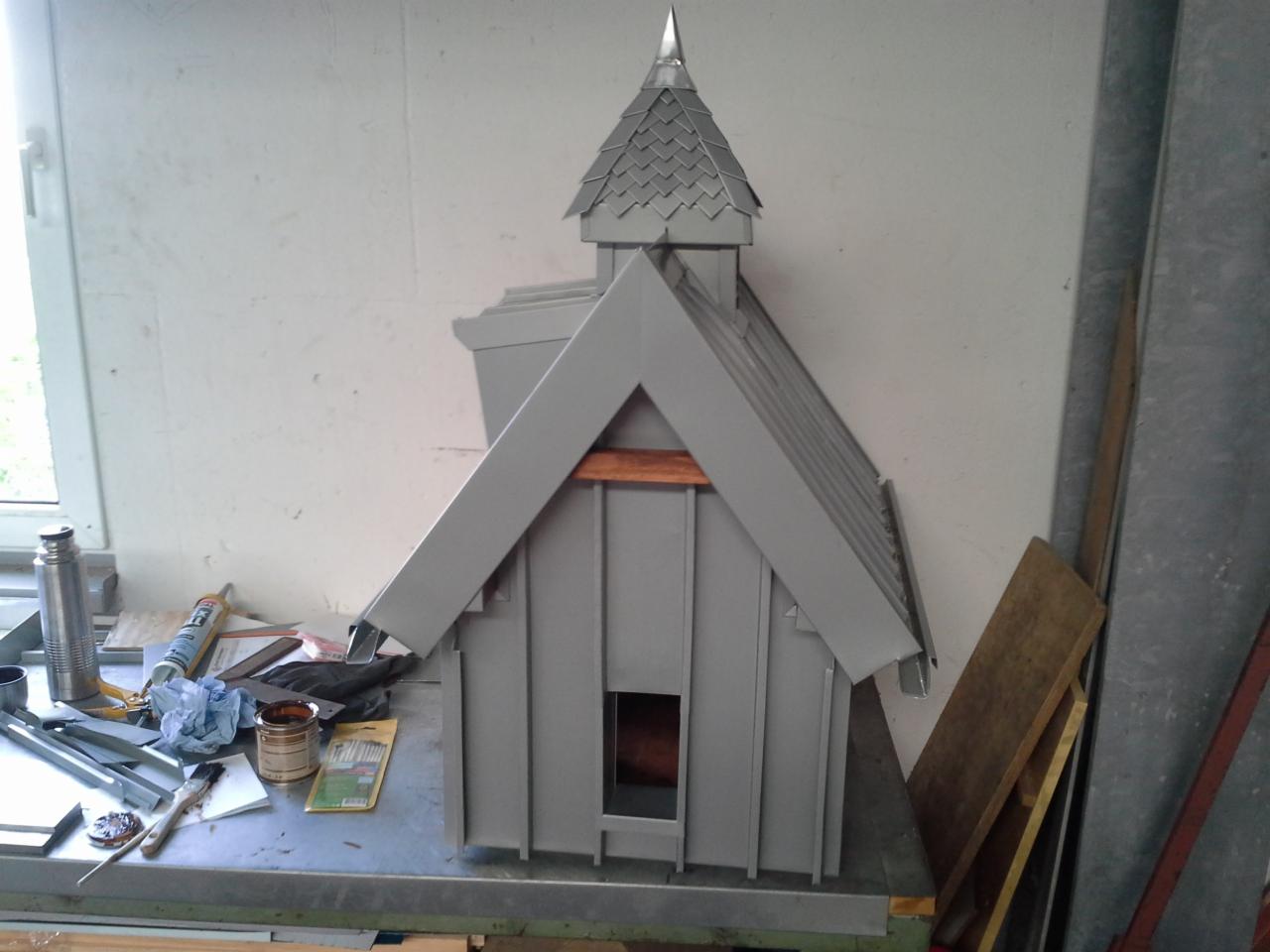 Szary model kapliczki lub domku z blachy z metalowym dachem łuskowym, wykonany na stole warsztatowym z narzędziami i materiałami.