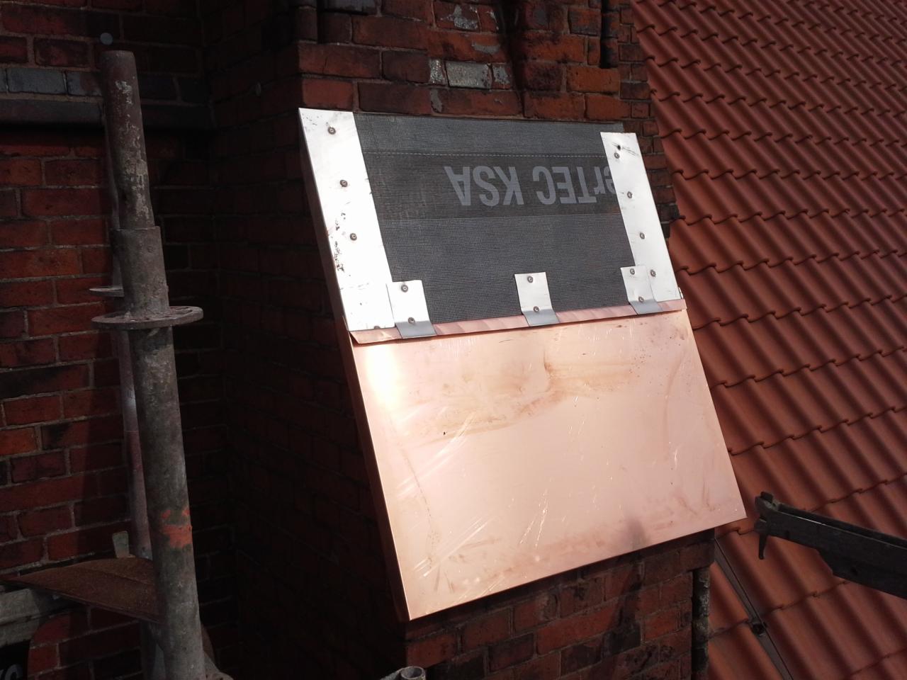 Miedziana osłona komina z elementami mocującymi na tle ceglanego komina i dachu z czerwonej dachówki.