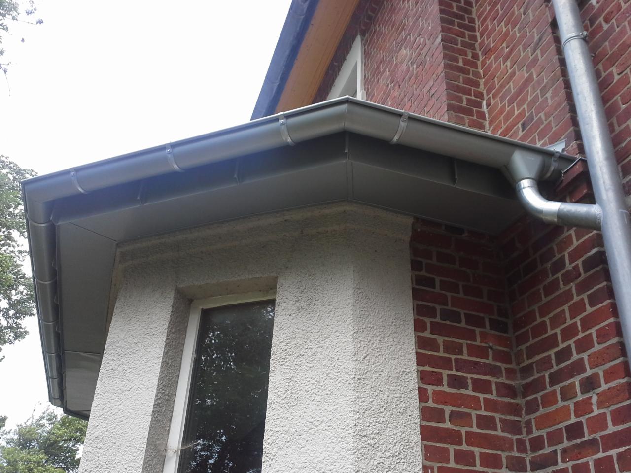 Szare orynnowanie budynku z cegły i tynkiem strukturalnym, widoczne mocowania i rura spustowa.