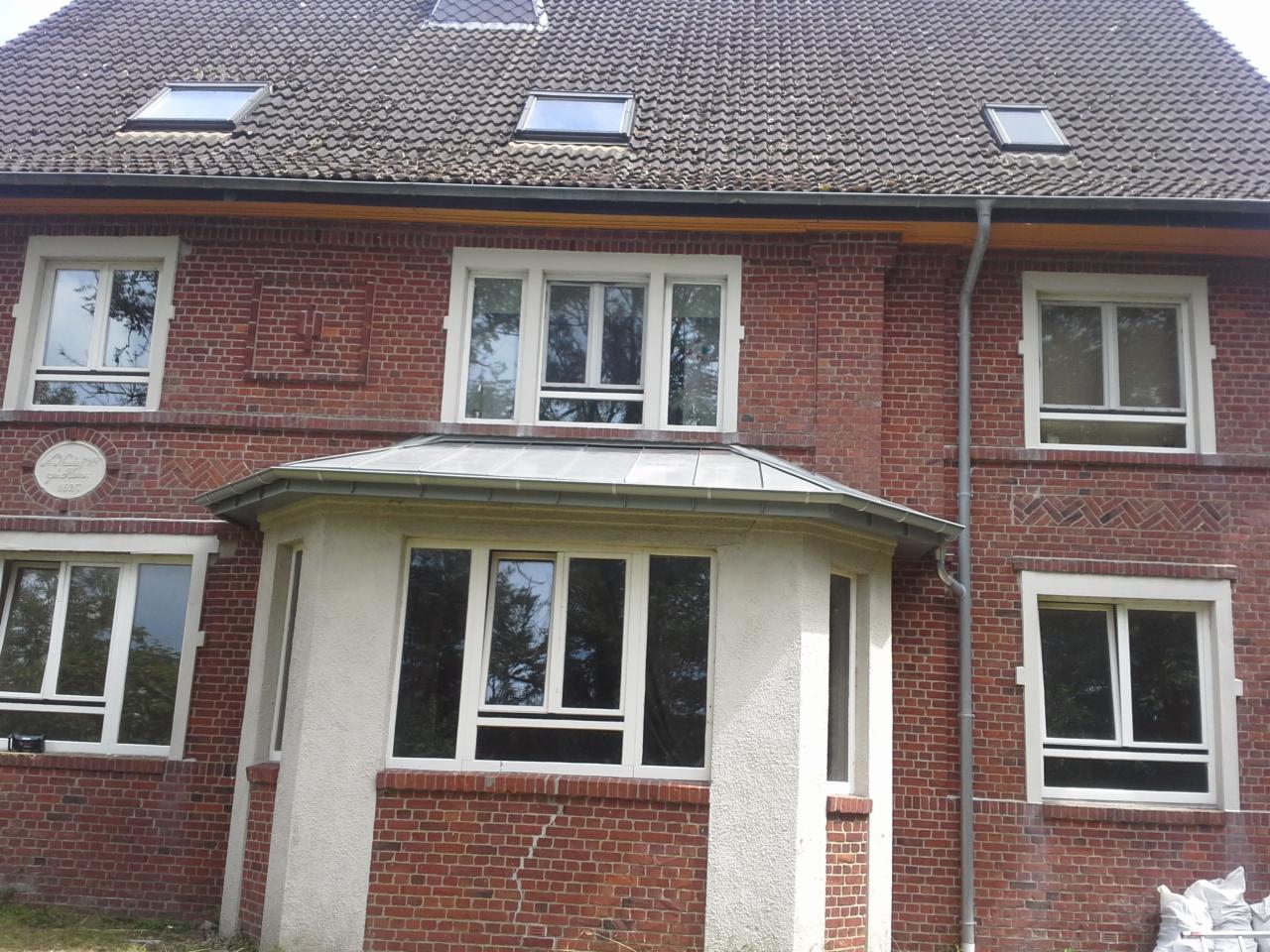 Ceglany budynek z szarym dachem, widoczne okna dachowe, rynna spustowa i fragment elewacji z datą 1897