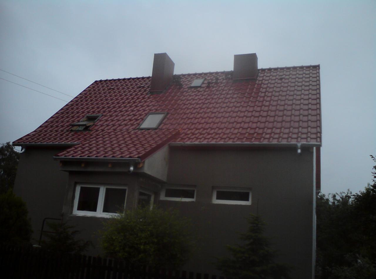 Dom jednorodzinny z nowym, czerwonym dachem z dachówki ceramicznej, widoczne kominy, okna dachowe i rynny, pochmurny dzień.