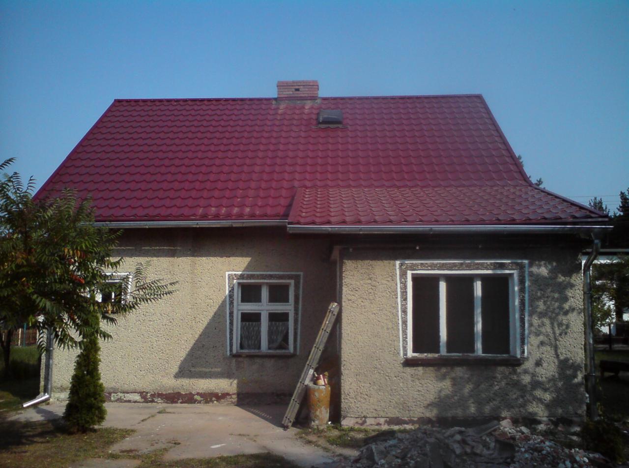 Dom jednorodzinny z nowym, czerwonym dachem z blachodachówki, widoczne rynny i komin, drabina oparta o ścianę, gruz pod oknem.