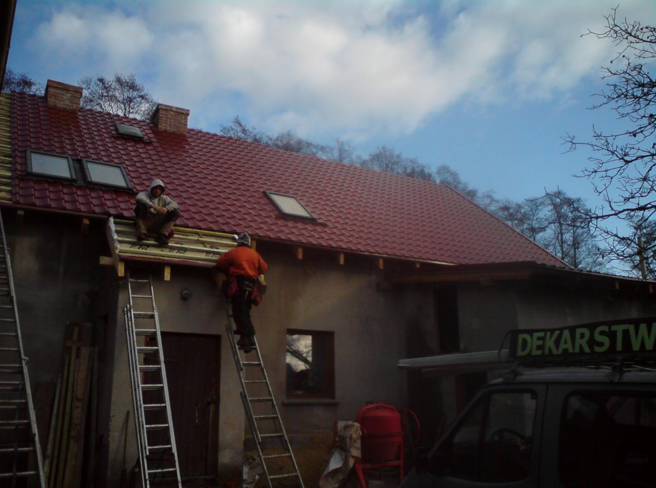 Dekarze montują dachówkę na dachu budynku z oknami dachowymi, jeden siedzi na dachu, drugi wchodzi po drabinie, widoczny samochód z napisem 'Dekarstwo'.