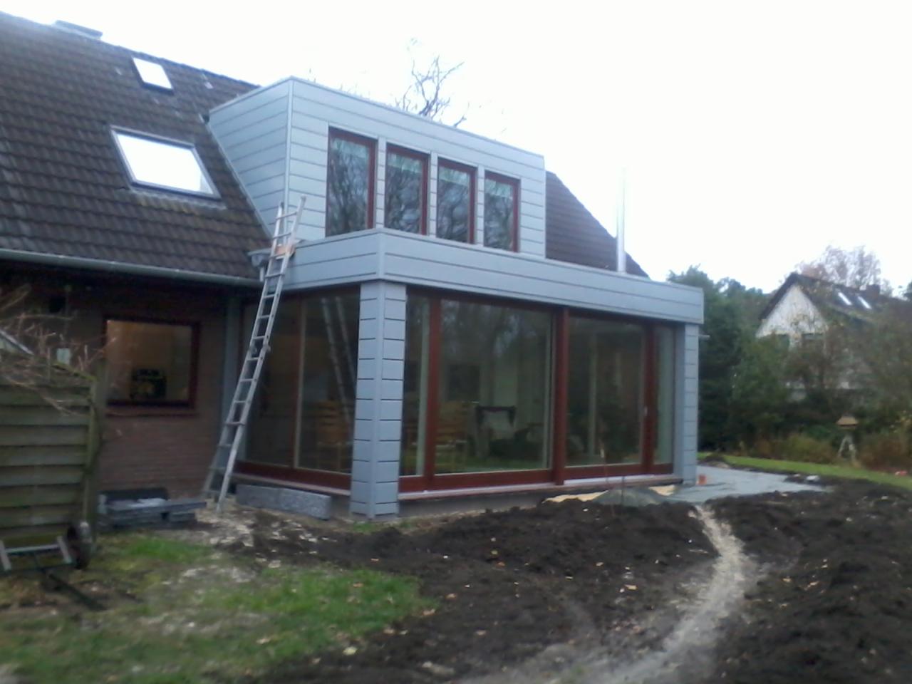 Dobudowa do domu z szarą elewacją, widoczne okna i drabina oparta o dach, teren wokół w trakcie prac ziemnych.