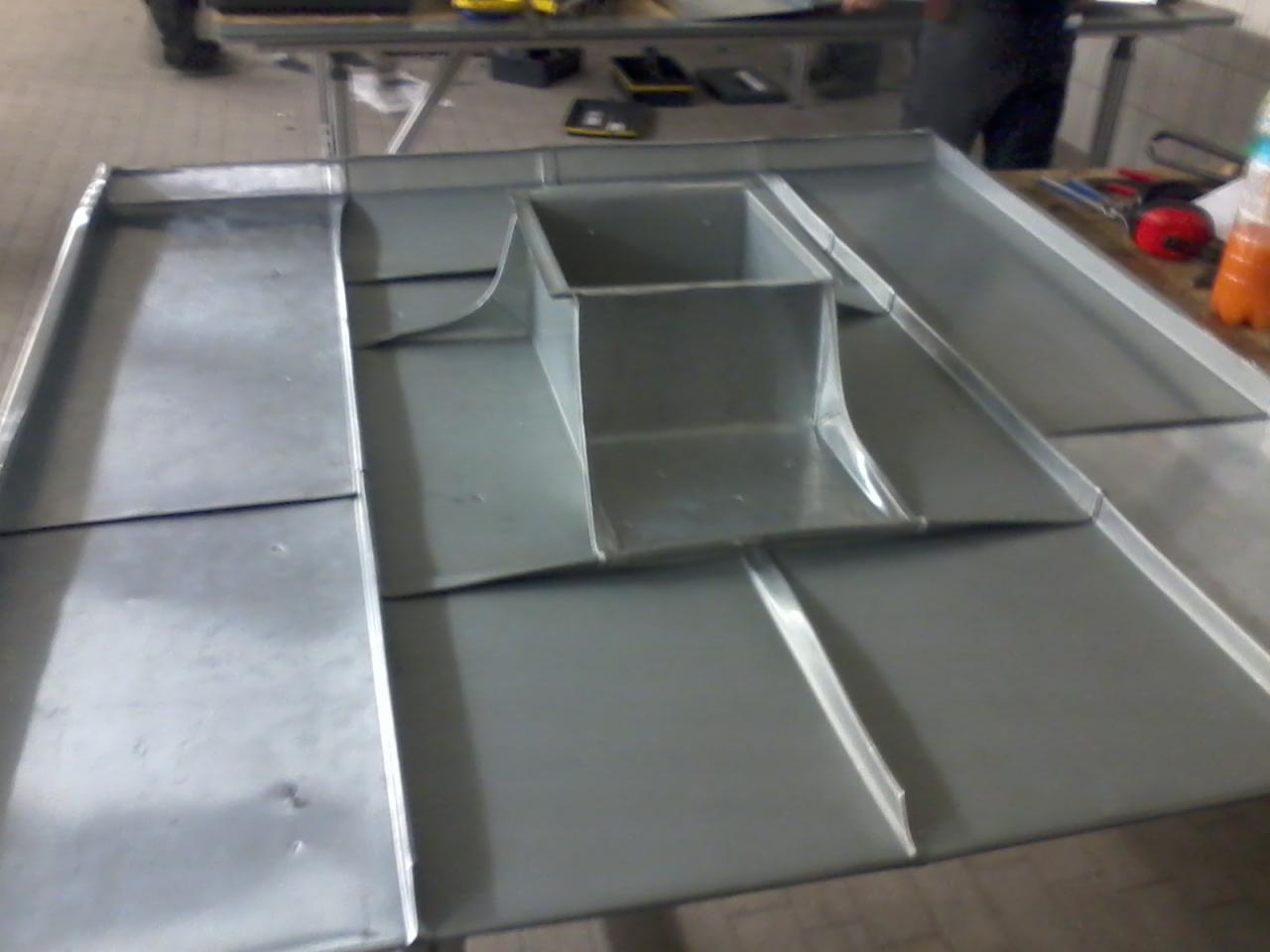 Szare, metalowe elementy przygotowane do montażu na dachu, widoczne połączenia i zagięcia blachy, fragment warsztatu w tle.