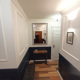 Usługi remontowo-budowlane Piotr Piosicki - Korytarz po remoncie: białe ściany z ciemnoniebieską lamperią, drewnopodobne płytki, lustro w ozdobnej ramie i mała konsola. Widoczne detale wykończenia.