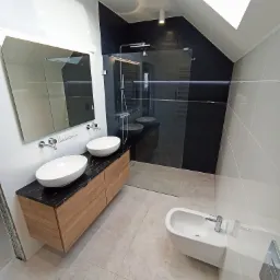 Nowoczesna łazienka na poddaszu z kabiną prysznicową typu walk-in, umywalkami nablatowymi na drewnianej szafce z czarnym blatem i oknem dachowym.