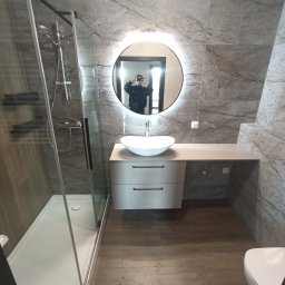 Usługi remontowo-budowlane Piotr Piosicki - Nowoczesna łazienka w szarościach: kabina prysznicowa ze szklanymi drzwiami, umywalka na szafce z szufladami, okrągłe lustro z podświetleniem LED i marmurowe płytki.