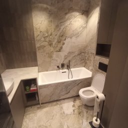 Usługi remontowo-budowlane Piotr Piosicki - Elegancka łazienka z wanną, umywalką i toaletą, wykończona płytkami imitującymi marmur. Nowoczesne oświetlenie punktowe i minimalistyczne meble w odcieniach szarości.