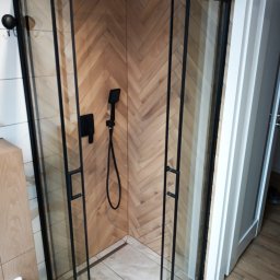 Usługi remontowo-budowlane Piotr Piosicki - Kabina prysznicowa z czarnymi profilami na tle ściany wyłożonej drewnopodobną płytką w jodełkę. Widoczna słuchawka prysznicowa i jasna podłoga.