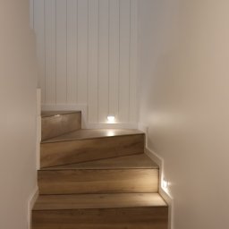 Usługi remontowo-budowlane Piotr Piosicki - Drewniane schody z oświetleniem punktowym wzdłuż ściany i panelami ściennymi. Nowoczesne wykończenie wnętrza domu, minimalistyczny design.