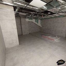 Usługi remontowo-budowlane Piotr Piosicki - Wnętrze w szarościach z ułożoną podłogą z płytek i fragmentem sufitu podwieszanego. Na podłodze leżą płyty gipsowe. Widoczne ślady po instalacjach.